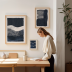 A woman is standing at a wooden desk, focused on reading a book, with two framed artworks hanging on the wall behind her.
