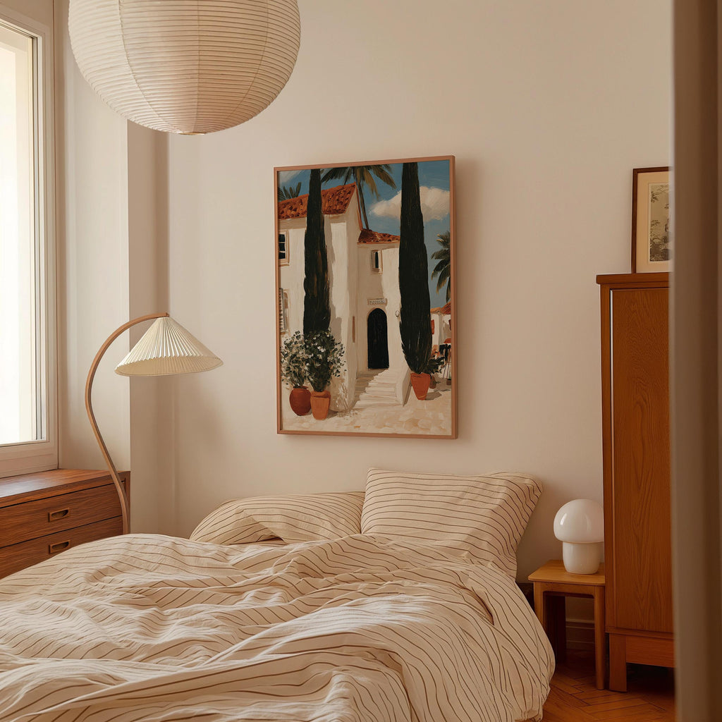 a cozy bedroom with a large bed, a wooden dresser, and a lamp. The bed has a striped comforter and a white pillow. Above the bed, there is a framed painting of a house with a palm tree. The room has a warm and inviting atmosphere, with natural light coming in from a window on the left side.