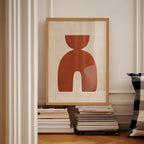 a framed abstract artwork with a bold red shape, displayed on a shelf among a stack of books.