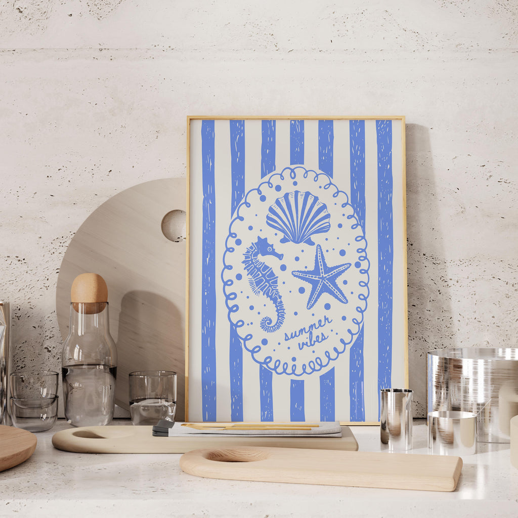 a framed artwork with a blue and white design depicting a seahorse, starfish, and seashell, along with the text "summer vibes" written around the edges. The artwork is displayed on a shelf, surrounded by various kitchen items such as a bottle, cups, and a cutting board.