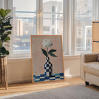 a large painting of a white flower in a checkered vase, placed on a wooden floor in front of a window overlooking a cityscape.