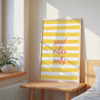 a framed poster with the text "good vibes only" in a yellow and white striped design, placed on a wooden table next to a potted plant.