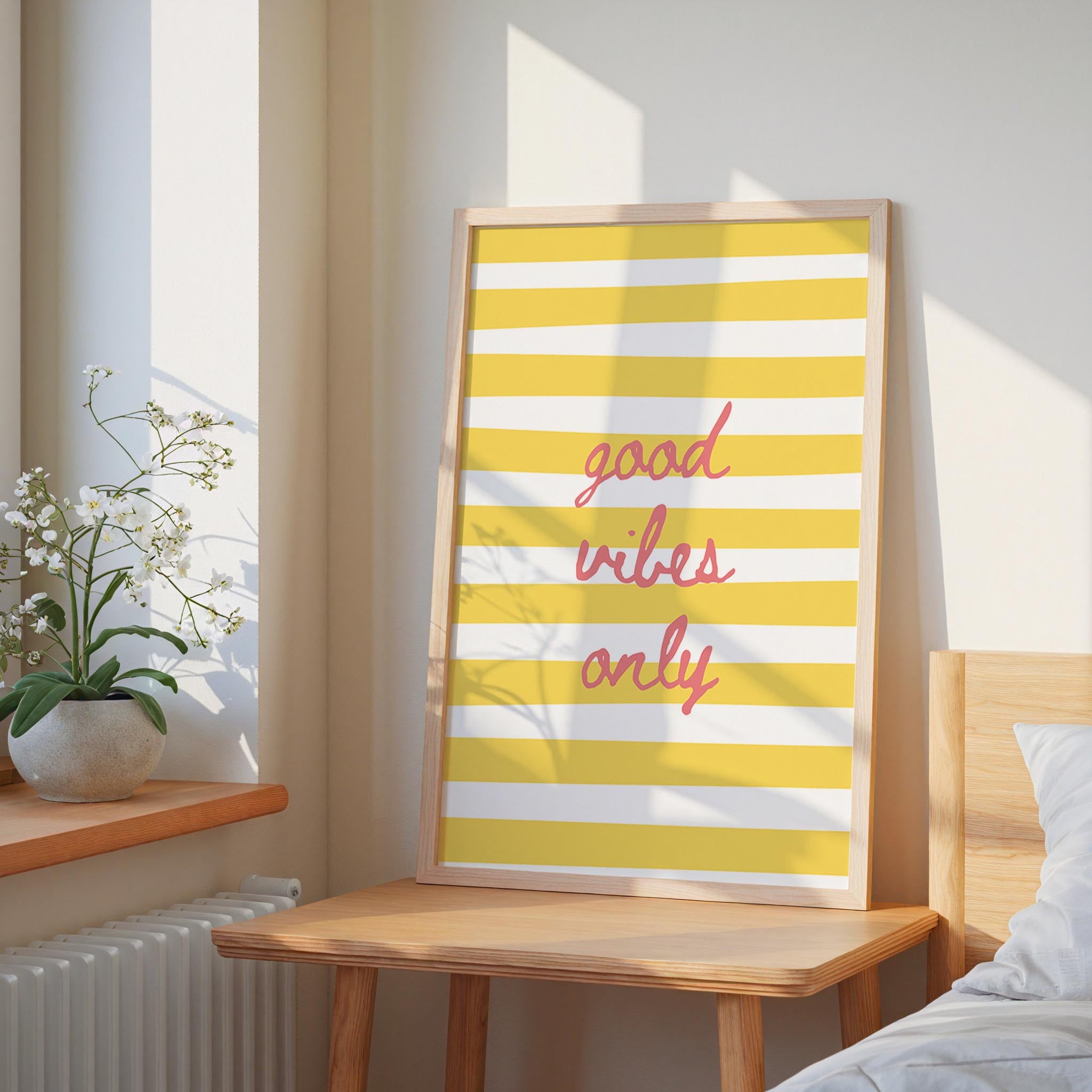 a framed poster with the text "good vibes only" in a yellow and white striped design, placed on a wooden table next to a potted plant.