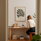 A woman is sitting at a wooden desk, surrounded by various objects, including a plant, books, and a framed leaf-like artwork on the wall.
