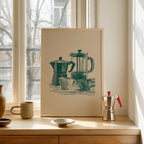a framed illustration of two coffee makers on a windowsill, with a small white bowl and a red coffee pot placed in front of the framed image.