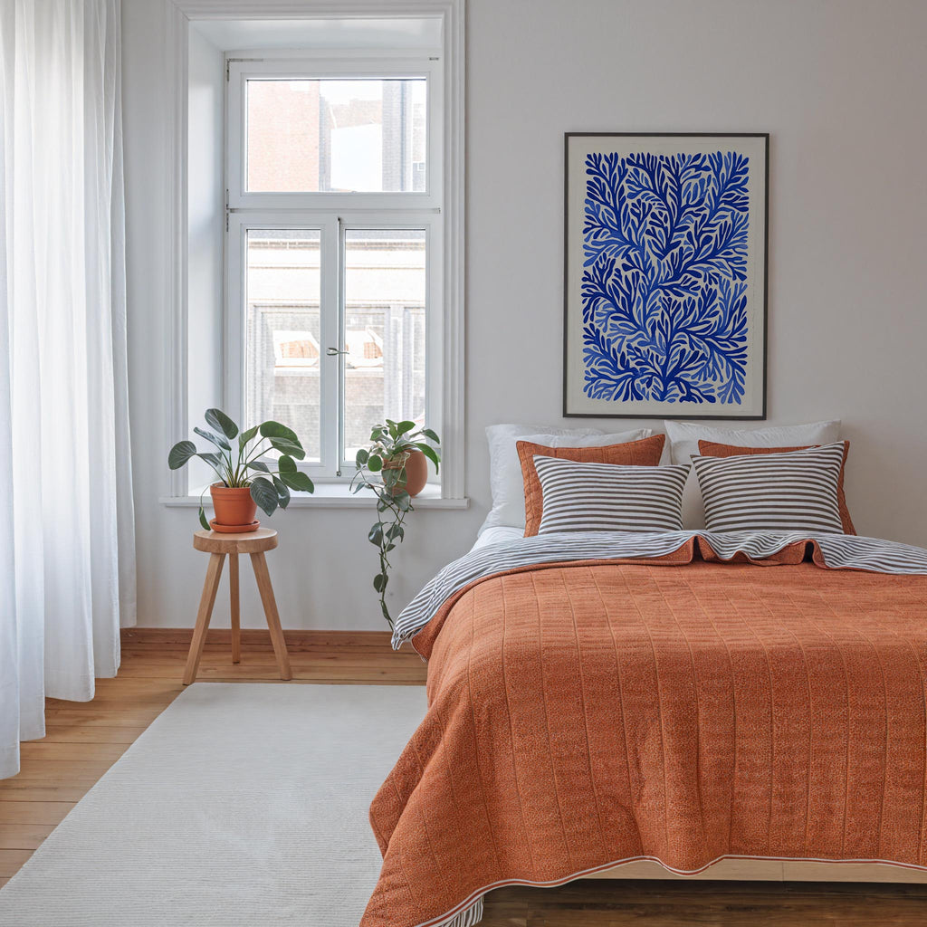 a cozy bedroom with a large bed, a wooden stool with a potted plant, and a framed artwork featuring a blue floral pattern on the wall.