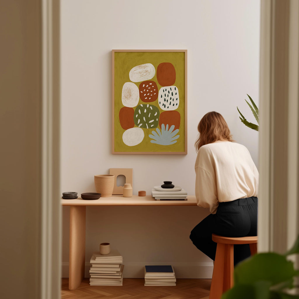 A woman is sitting at a wooden desk, surrounded by various objects, including a stack of books, a plant, and a framed abstract art piece on the wall.