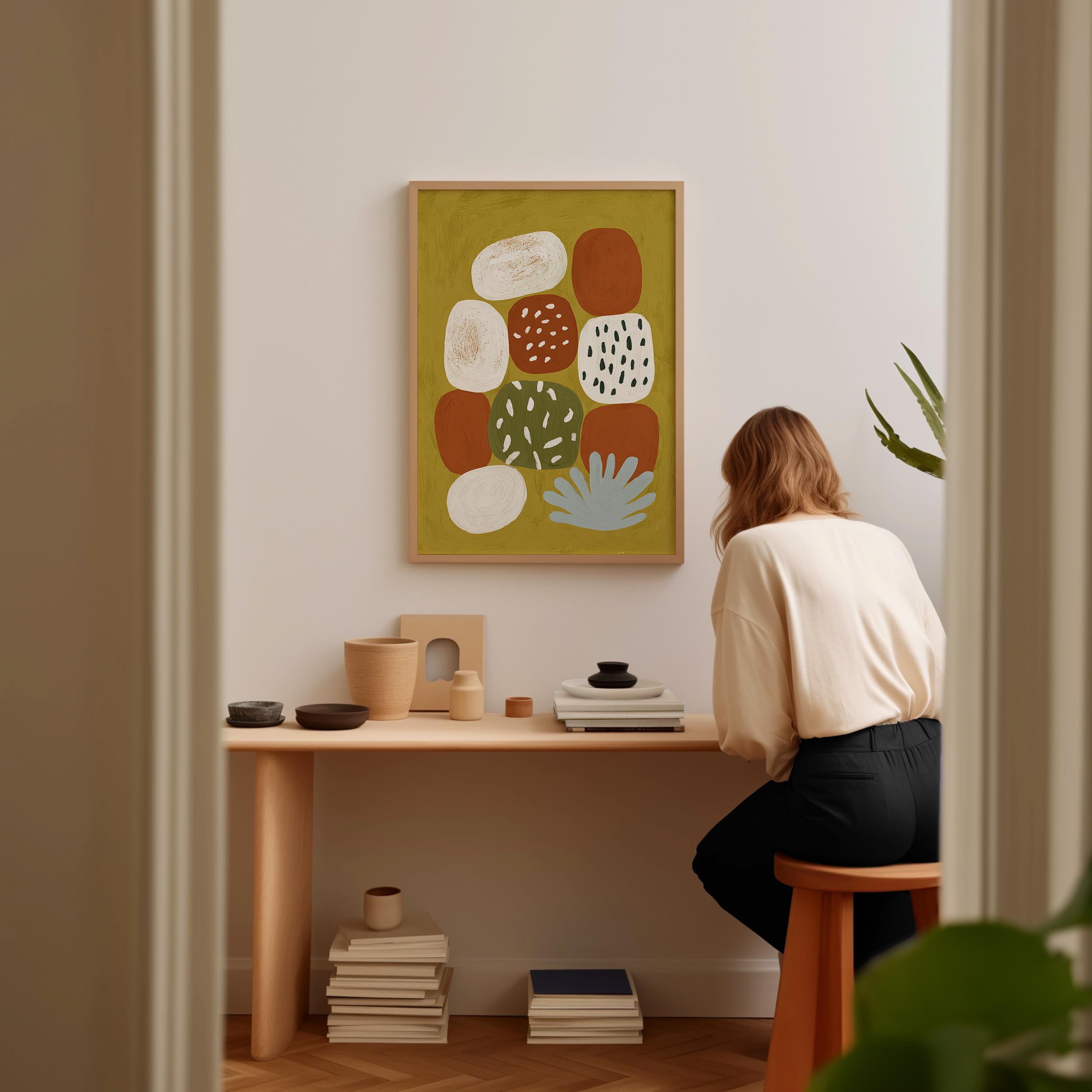 A woman is sitting at a wooden desk, surrounded by various objects, including a stack of books, a plant, and a framed abstract art piece on the wall.