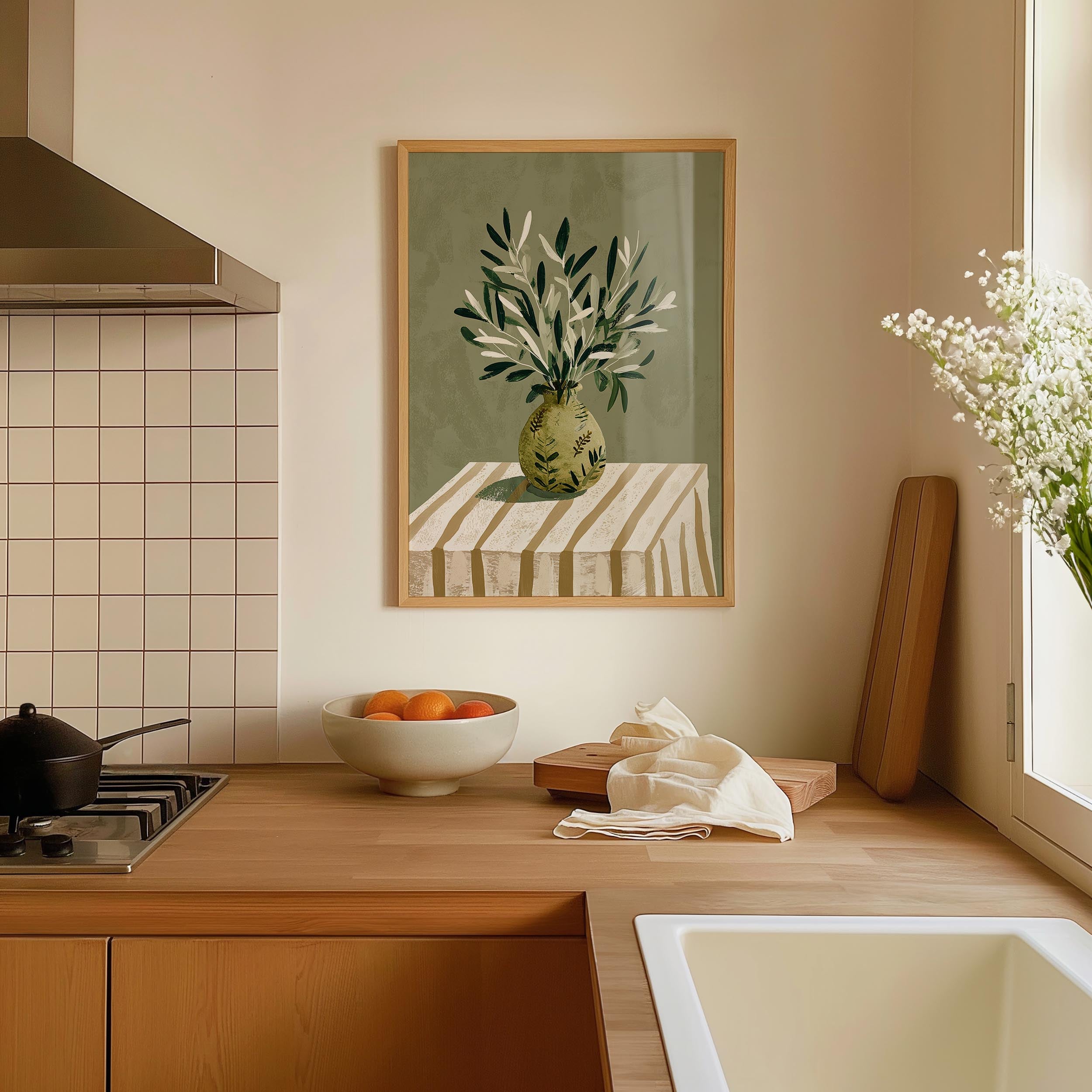 a kitchen counter with a bowl of fruit, a vase of flowers, and a wooden cutting board. The counter is made of wood and has a sink and a stove in the background. A framed painting of a vase of flowers is hanging on the wall above the counter.