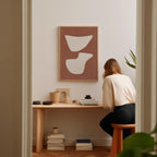 A woman is sitting at a wooden desk, surrounded by various objects, including a framed abstract artwork, a stack of books, and a plant.