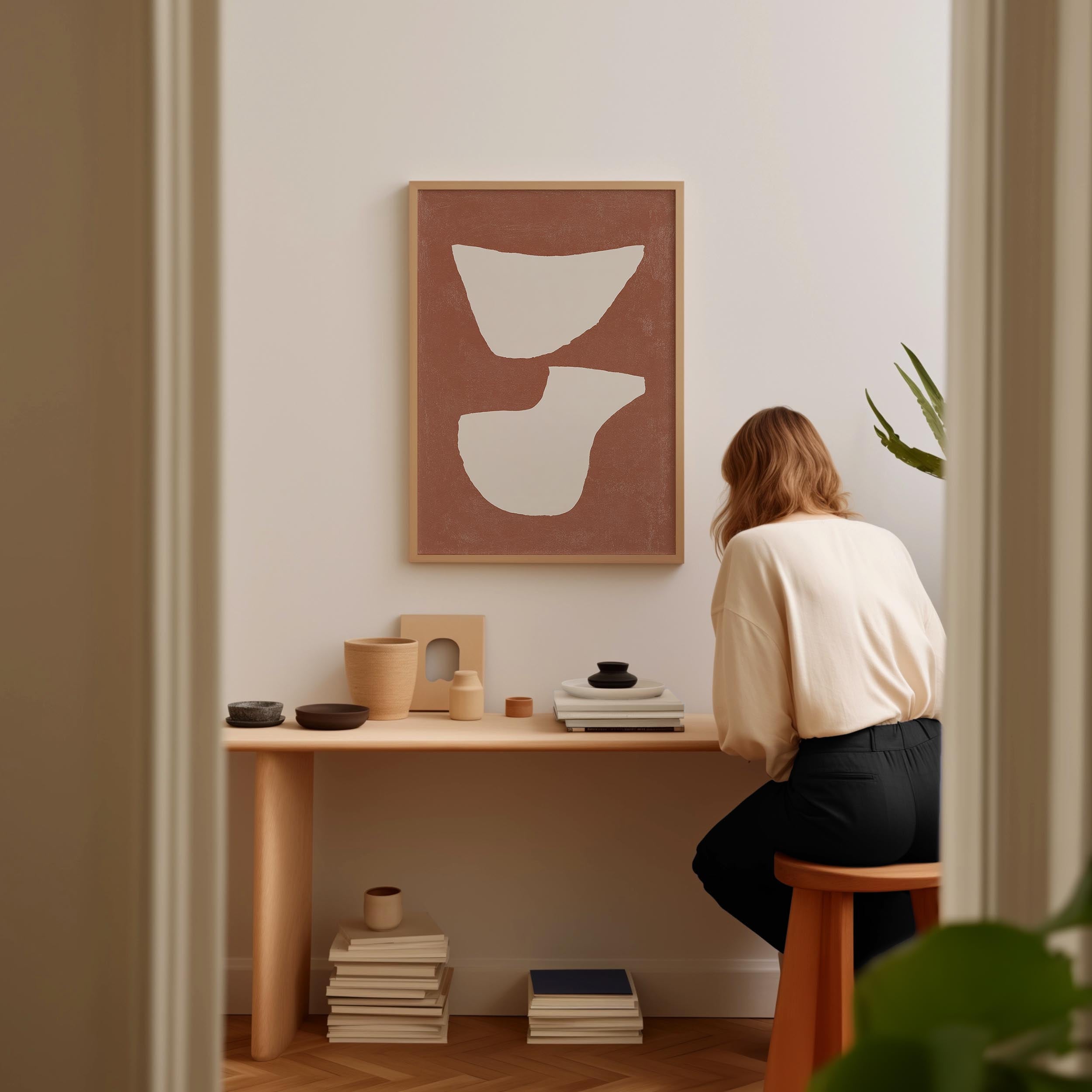 A woman is sitting at a wooden desk, surrounded by various objects, including a framed abstract artwork, a stack of books, and a plant.