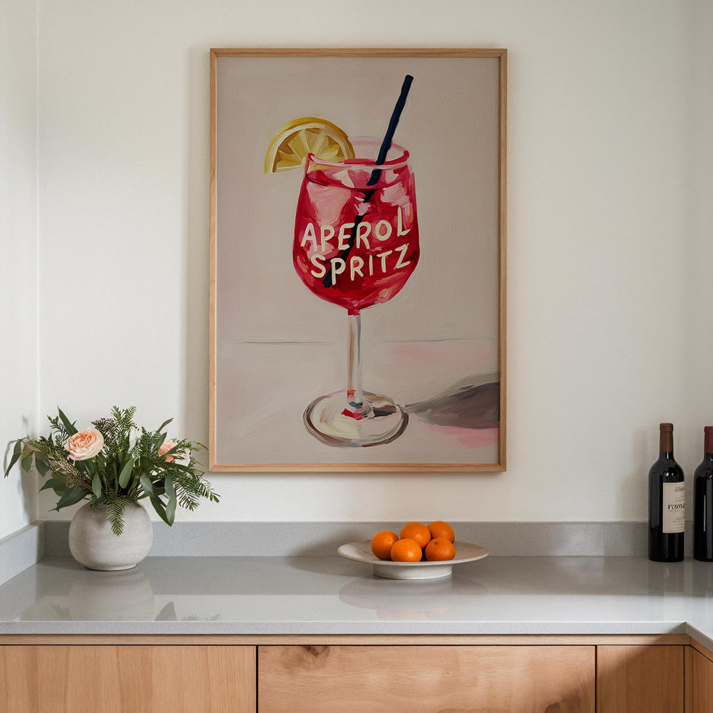 a framed painting of a red drink with a lemon slice and a straw, hanging on a wall above a counter with a vase of flowers and a plate of oranges.