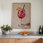 a framed painting of a red drink with a lemon slice and a straw, hanging on a wall above a counter with a vase of flowers and a plate of oranges.