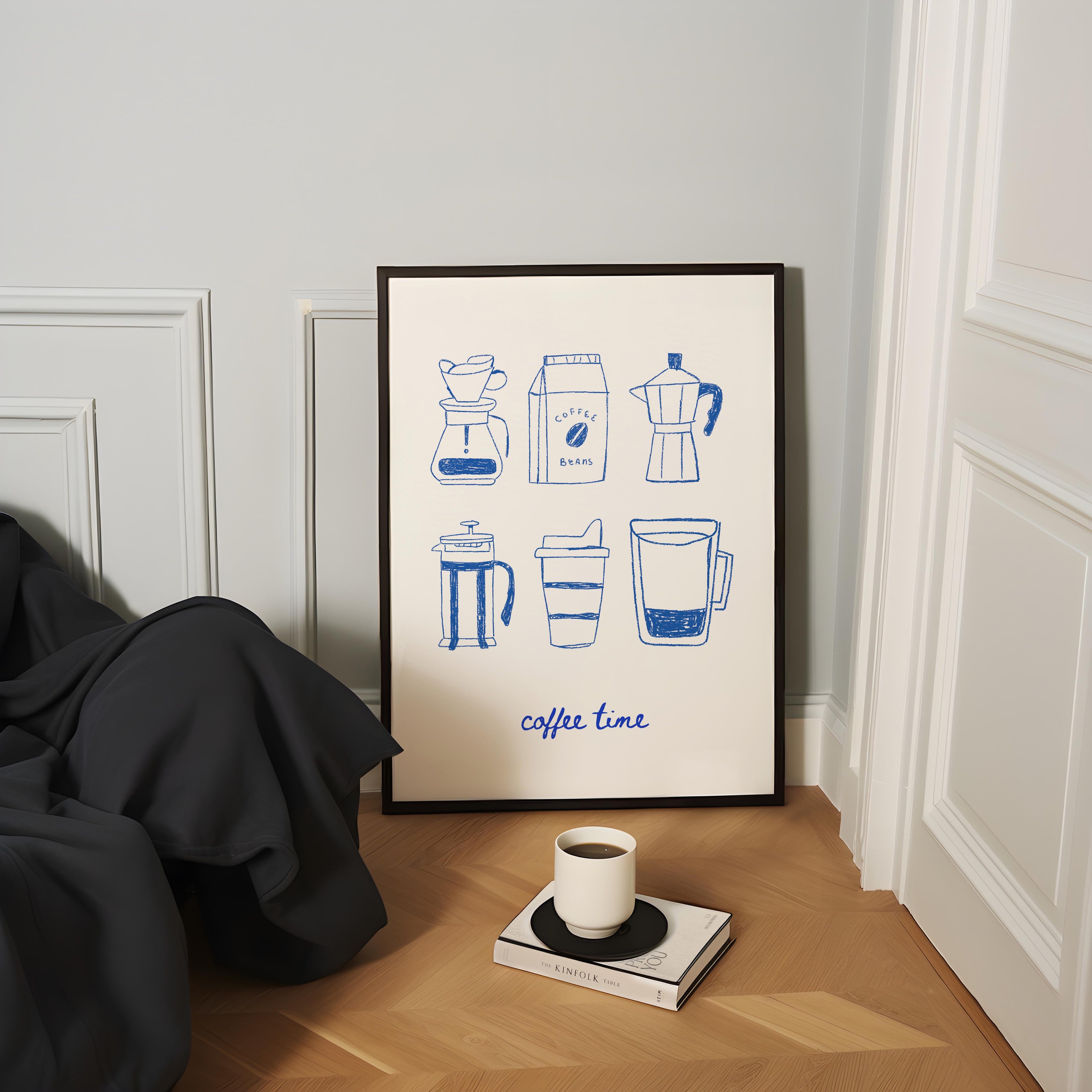 a framed poster with illustrations of coffee-making equipment, including a French press, coffee maker, and a thermos, along with the phrase "coffee time" written below. A cup of coffee is placed on a small table next to the poster, and there is a book and a blanket on the floor in the foreground.