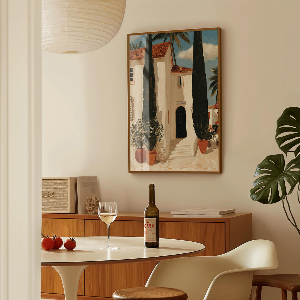 a dining area with a table set for two, a bottle of wine, and a framed painting of a house with a palm tree in the background.