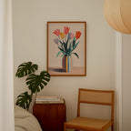 a cozy corner of a room with a wooden chair, a wooden side table, a framed painting of a vase with colorful tulips, and a potted plant.