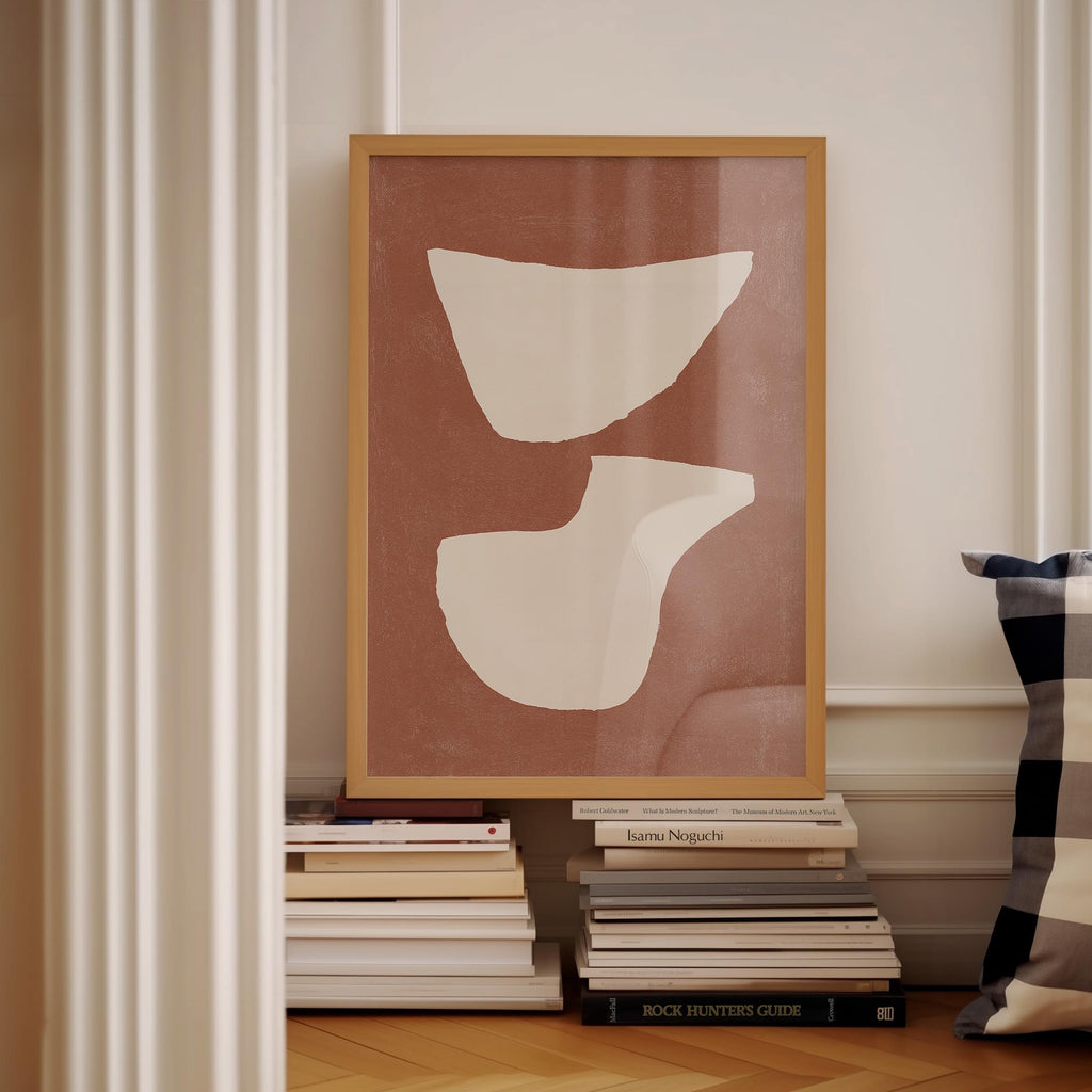 a framed abstract artwork with a brown background and white shapes, displayed on a shelf among a stack of books.