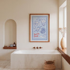 a bathroom with a large framed poster of a blue and white illustration hanging on the wall above a white bathtub. The poster depicts a seashell and a rose, adding a coastal touch to the room. The bathroom also includes a window, a shelf with toiletries, and a woven basket, creating a cozy and inviting atmosphere.