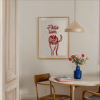 a dining area with a table, chairs, and a framed poster on the wall. The poster has a red and white design with the text "Pasta lover" written in cursive. There is also a vase of flowers on the table, adding a touch of color and life to the scene.