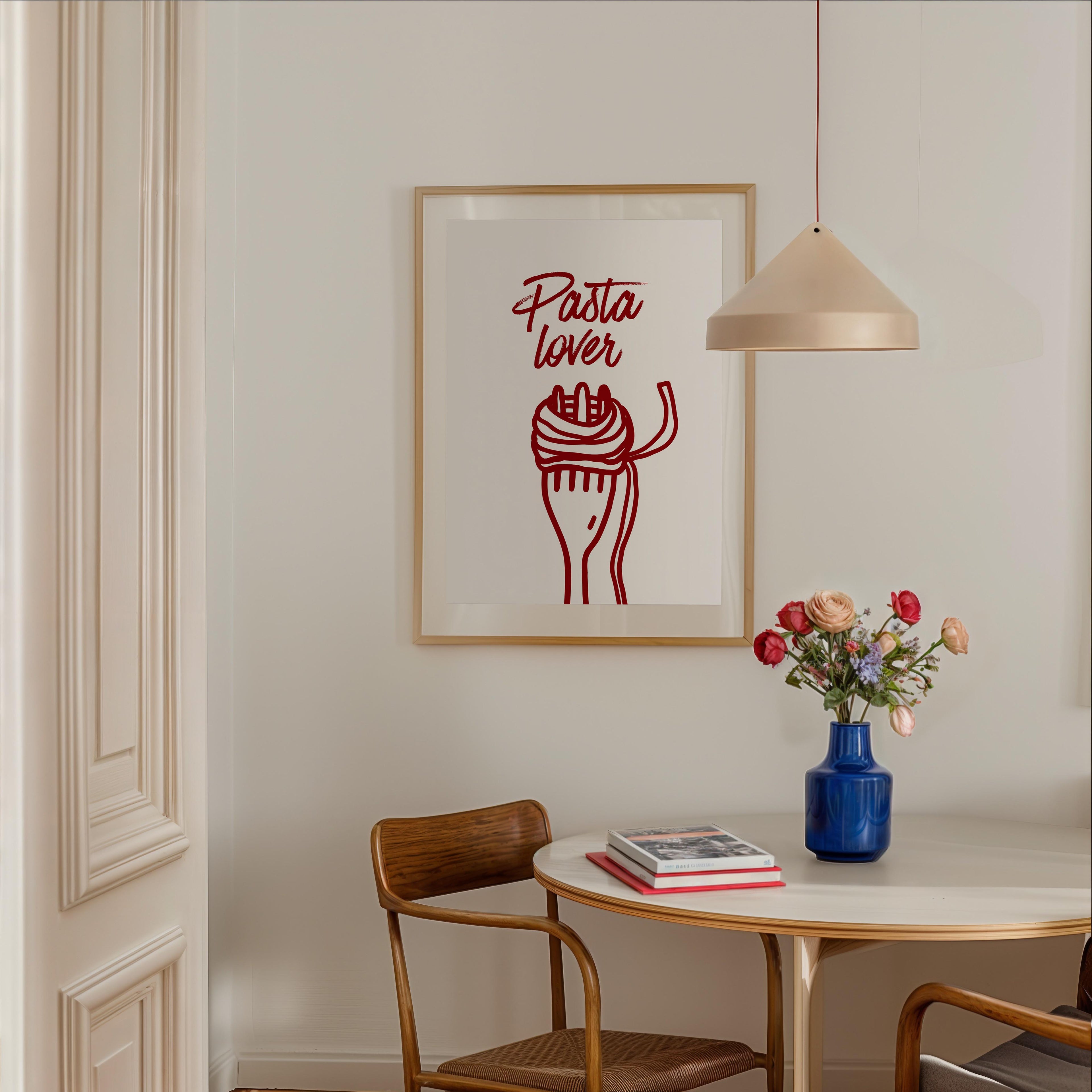 a dining area with a table, chairs, and a framed poster on the wall. The poster has a red and white design with the text "Pasta lover" written in cursive. There is also a vase of flowers on the table, adding a touch of color and life to the scene.