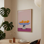 a minimalist dining area with a table, a vase of flowers, and a framed artwork depicting a bowl of fruit on a pink background.