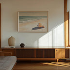 a serene beach scene with a framed painting hanging on the wall above a wooden cabinet or entertainment center. The painting depicts a tranquil beach with a single shell resting on the sand. The cabinet has a vase and a book on top, adding a touch of personalization to the space. The room is bathed in natural light, creating a warm and inviting atmosphere.