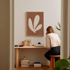 A woman is sitting at a wooden desk, surrounded by various objects, including a plant, books, and a framed abstract art piece.