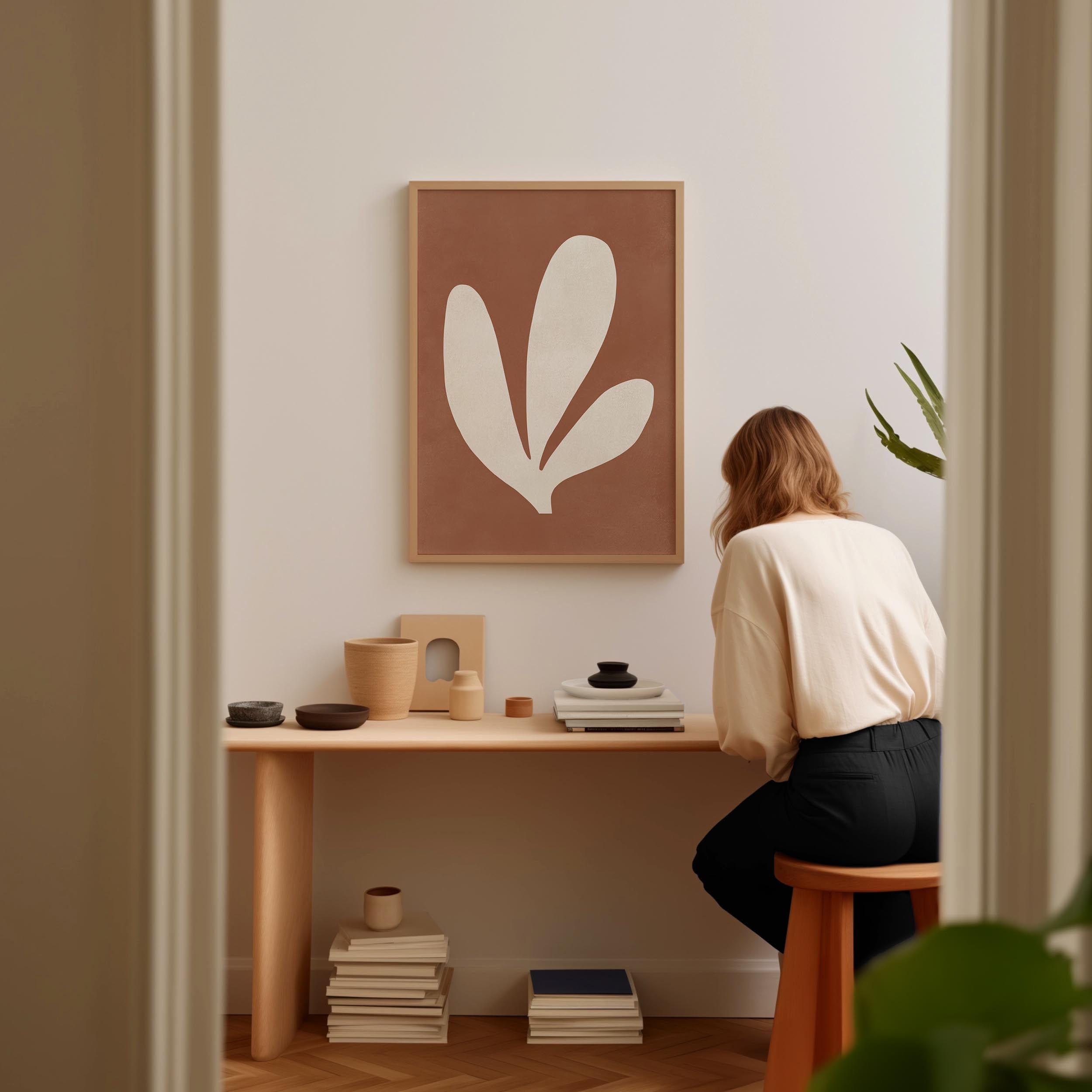 A woman is sitting at a wooden desk, surrounded by various objects, including a plant, books, and a framed abstract art piece.