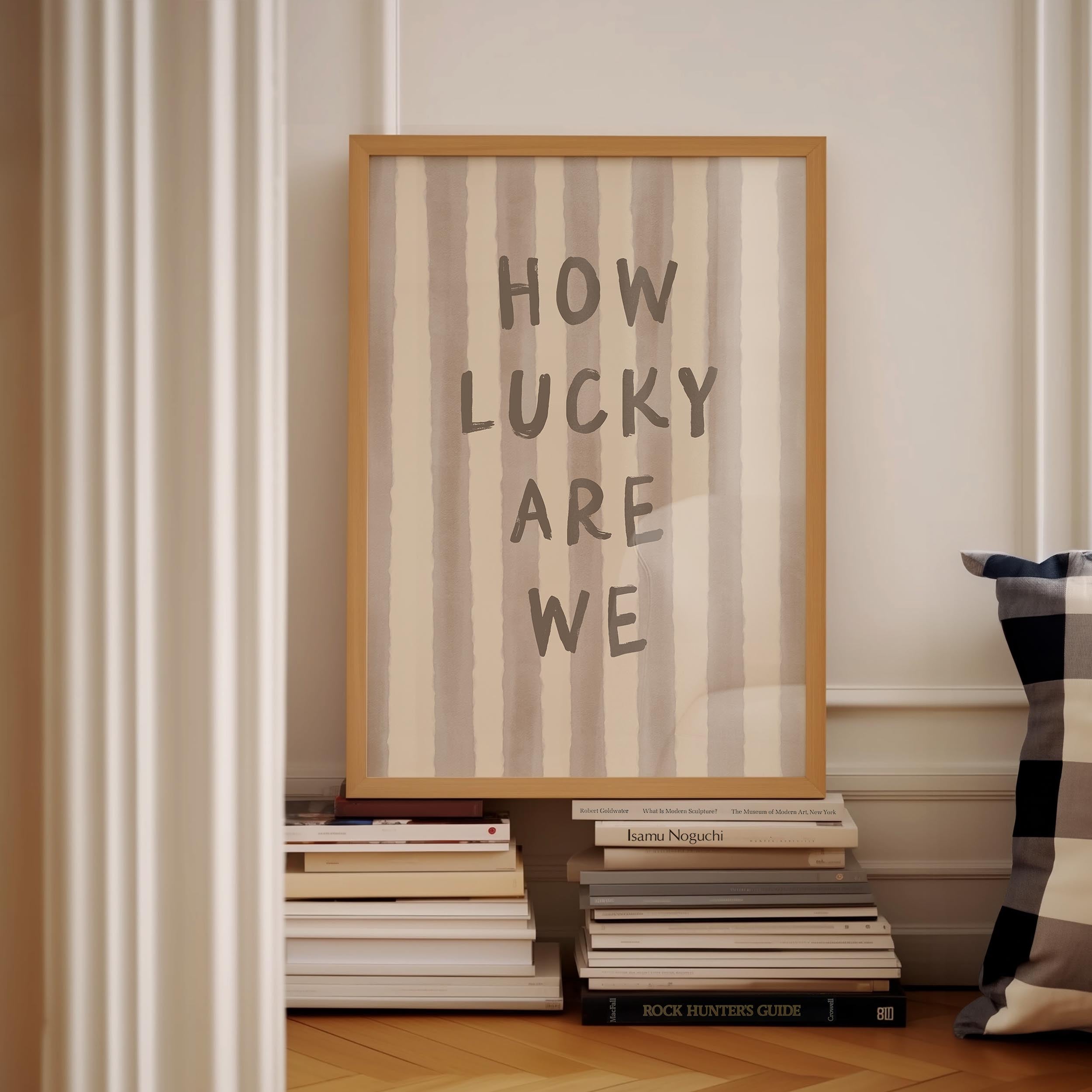 a framed poster with the text "HOW LUCKY ARE WE" displayed on it, placed on a shelf next to a stack of books.