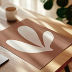 a brown paper with a white leaf-like design on it, placed on a wooden surface. In the background, there is a white cup and a plant, creating a cozy and natural atmosphere.