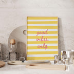 a yellow and white striped poster with the text "good vibes only" displayed on it, placed on a countertop with various kitchen items such as a bottle, cups, and a cutting board.