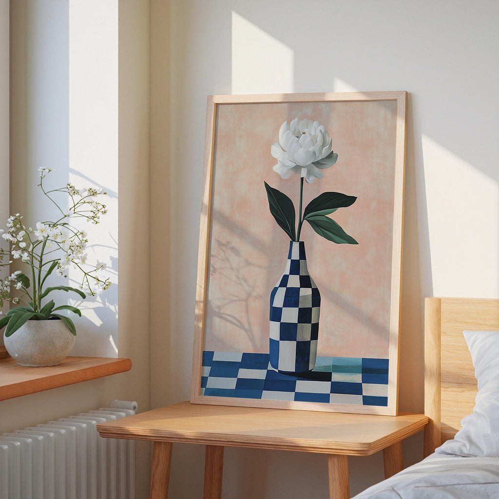 a framed painting of a white flower in a blue and white checkered vase, placed on a wooden table in a bedroom setting.