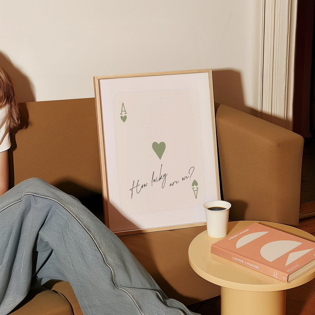 a person sitting on a couch with a framed poster of a playing card with a heart and the text "How lucky are we?" next to a small table with a book and a cup of coffee.