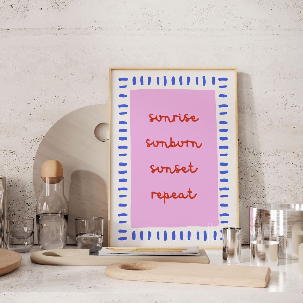 a pink framed poster with the words "sunrise, sunset, repeat" written on it, placed on a countertop next to various kitchen items such as a bottle, cups, and a cutting board.