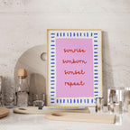a pink framed poster with the words "sunrise, sunset, repeat" written on it, placed on a countertop next to various kitchen items such as a bottle, cups, and a cutting board.