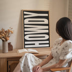 A woman in a white dress is sitting at a desk with an open book and a vase of flowers, with a large black and white sign that says "HOWDY" in the background.