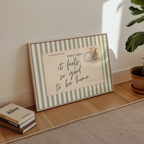 A framed postcard with a handwritten message sits on a wooden floor next to a stack of books and a potted plant.