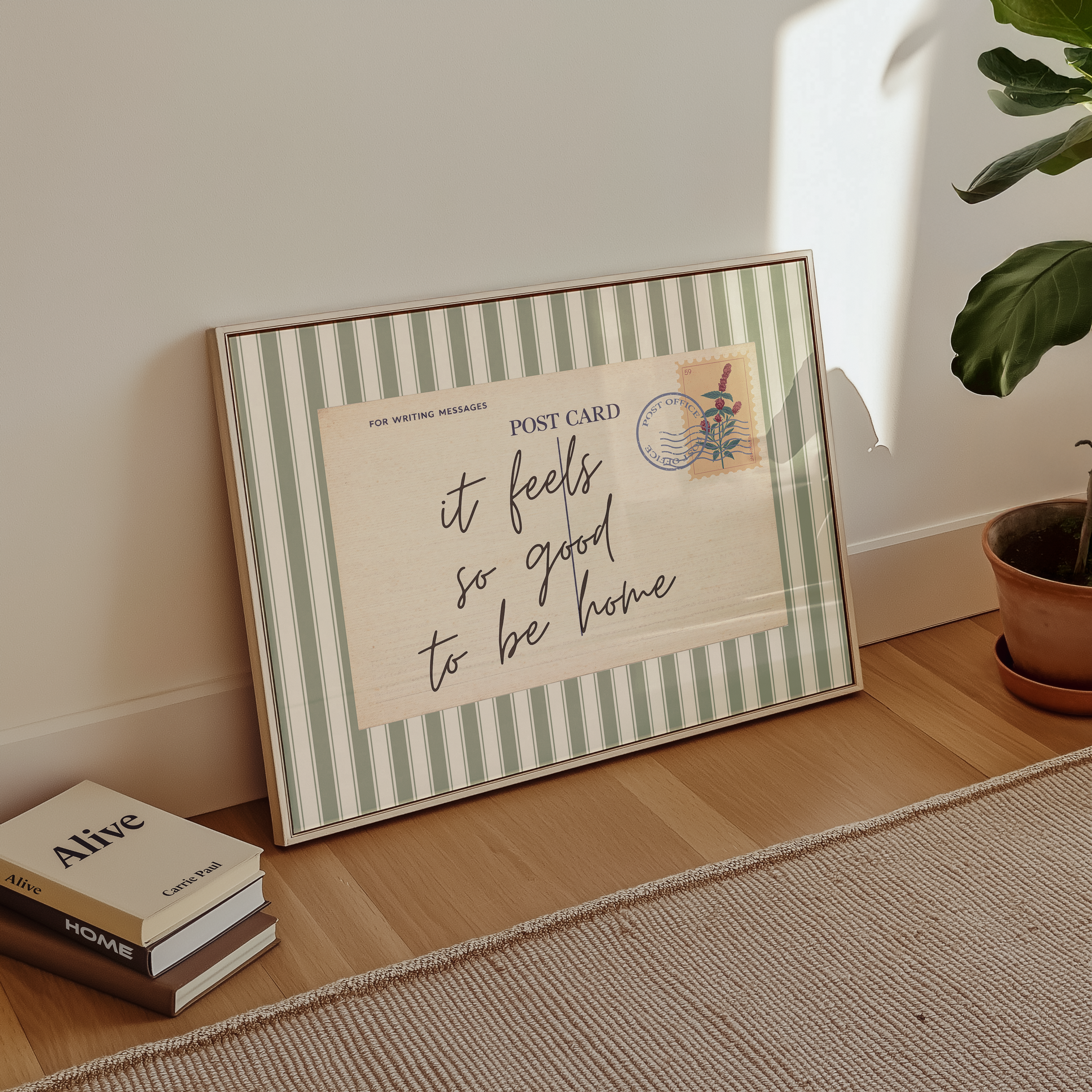 A framed postcard with a handwritten message sits on a wooden floor next to a stack of books and a potted plant.