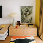 a cozy corner of a room with a wooden dresser, a lamp, a framed painting of a cat, and a stack of books.