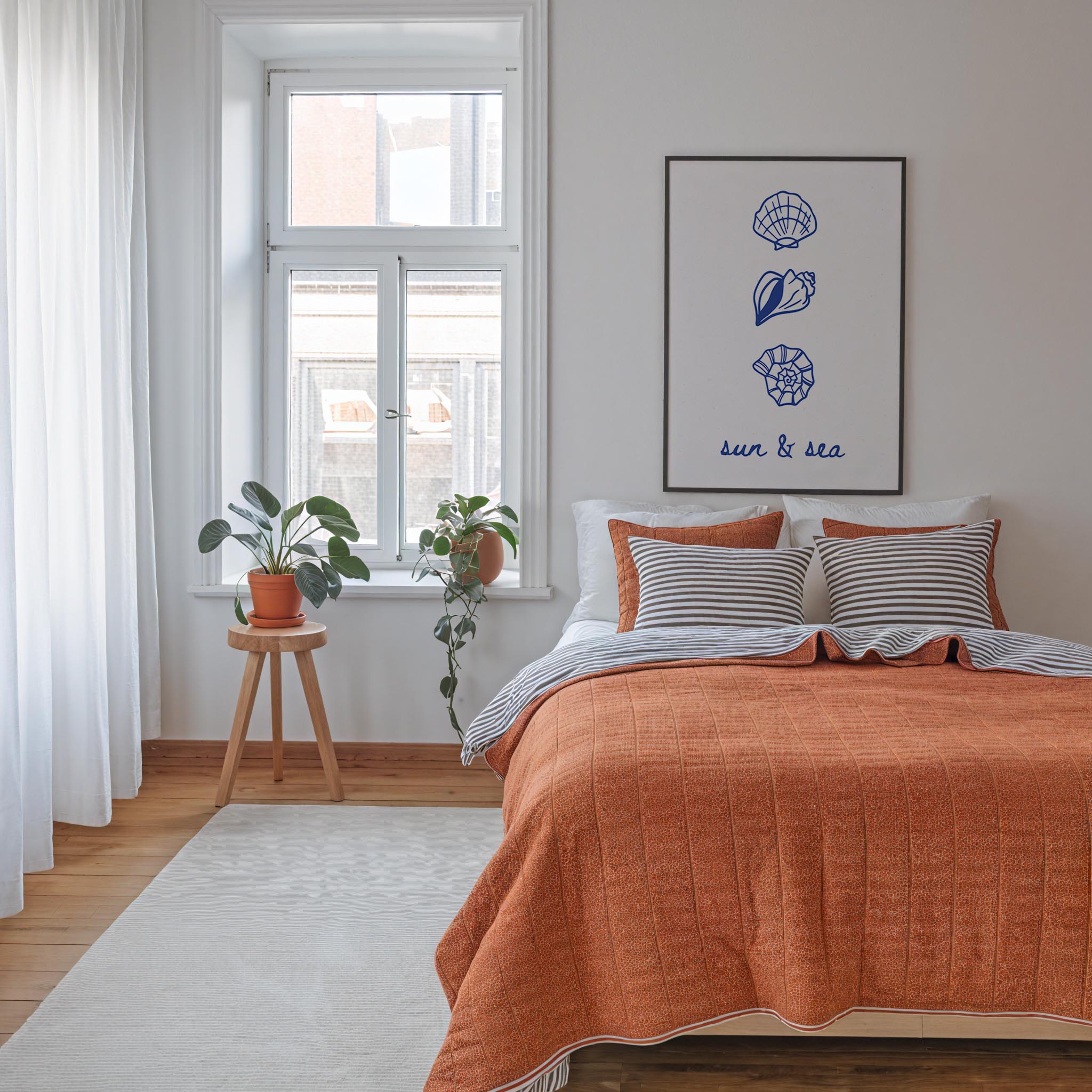 a cozy bedroom with a large bed, a window with a view of the outside, and a framed artwork featuring seashells and the words "sun & sea".