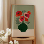 a framed print of a vase with red flowers on a green background, placed on a wooden chair.
