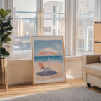 a large framed painting depicting a beach scene with a beach umbrella and a lounge chair, placed on a wooden floor in a room with large windows overlooking a cityscape.