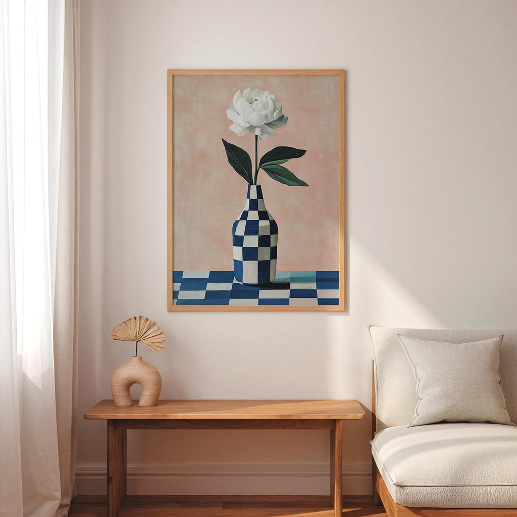 a living room with a wooden table, a white vase with a single white flower, and a beige sofa. Above the table, there is a framed painting of a white flower in a checkered vase.