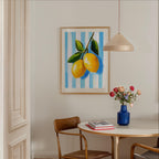 a dining area with a table, chairs, and a framed painting of lemons hanging on the wall. The table has a vase of flowers and a stack of books on it.