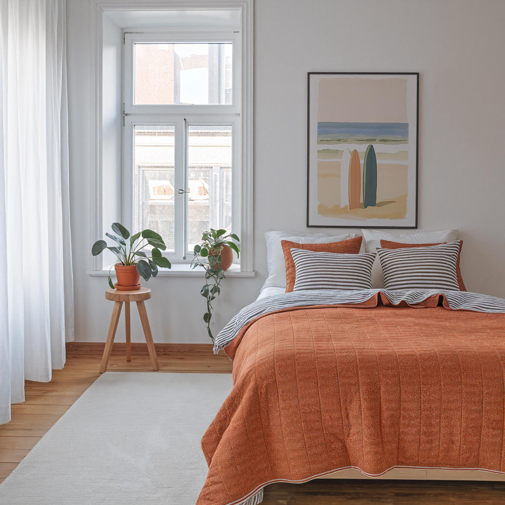 a cozy bedroom with a large bed, a framed artwork of surfboards on the wall, and a small table with a potted plant. The room has a warm and inviting atmosphere, with a neutral color palette and natural elements.