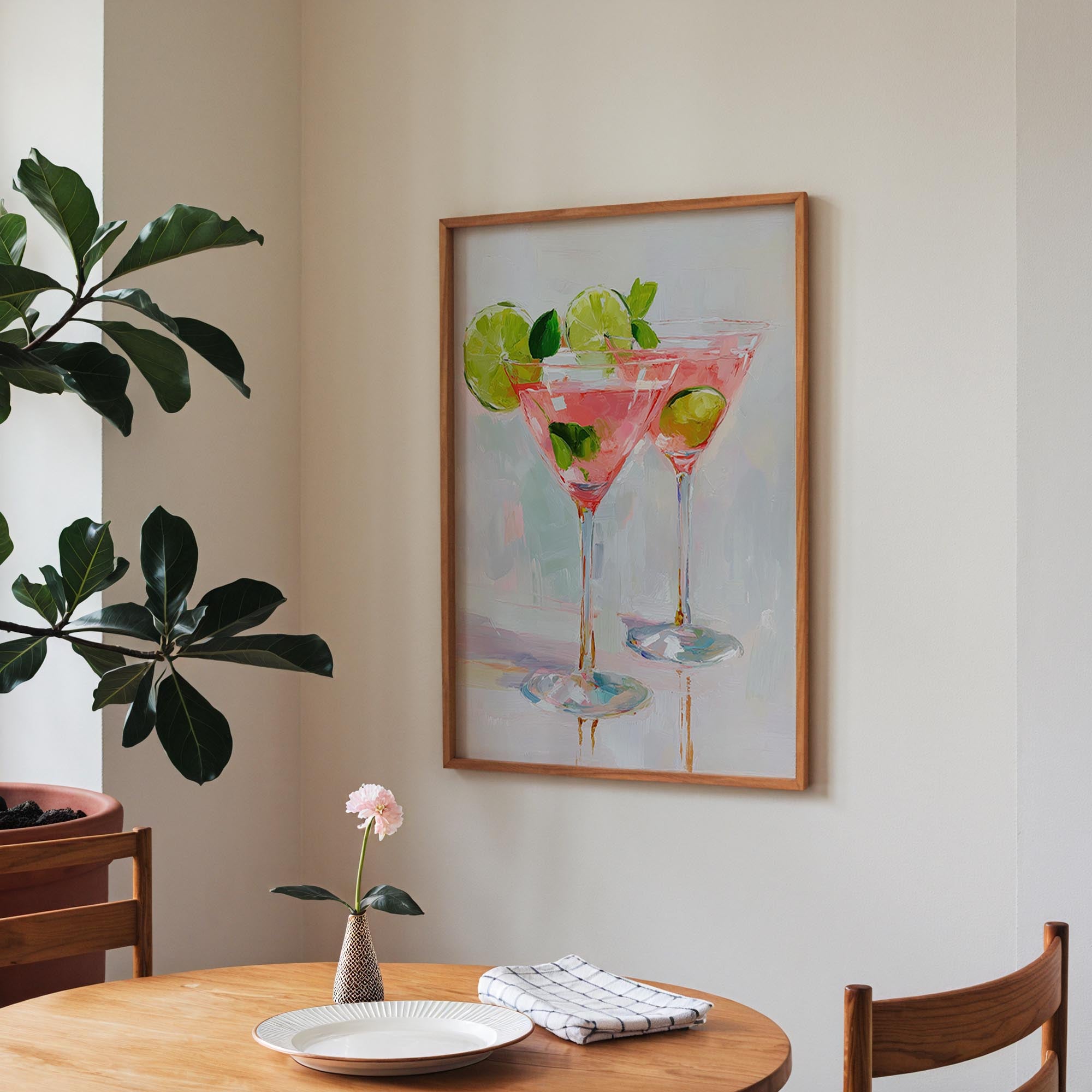 a framed painting of two martini glasses with lime slices, hanging on a wall above a wooden table with a vase of pink flowers and a plate with a white napkin.