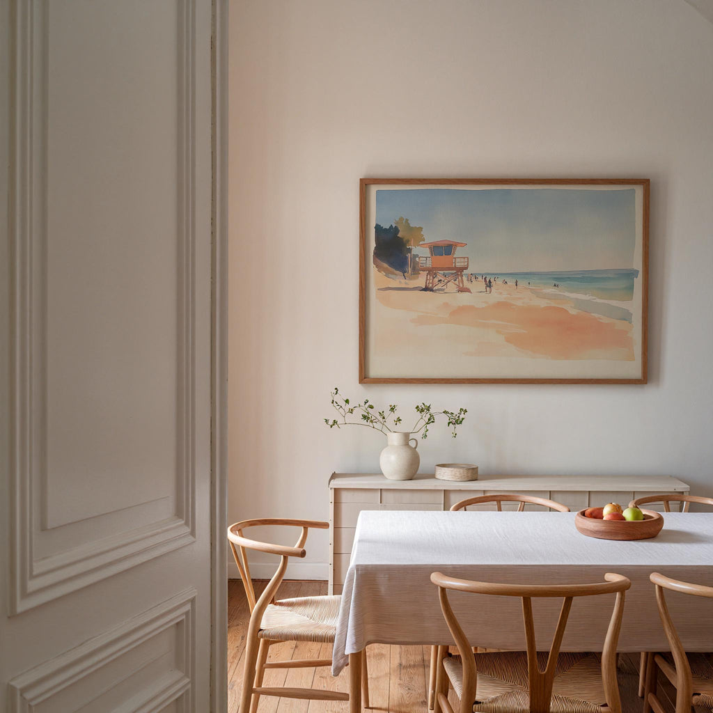 a dining room with a white table and chairs, a framed painting of a beach scene hanging on the wall, and a vase of flowers on the table.