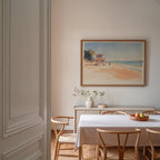 a dining room with a white table and chairs, a framed painting of a beach scene hanging on the wall, and a vase of flowers on the table.