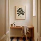 a cozy corner of a room with a wooden desk, a framed abstract leaf design, and a small plant on the desk. The room has a checkered floor and a window, creating a warm and inviting atmosphere.