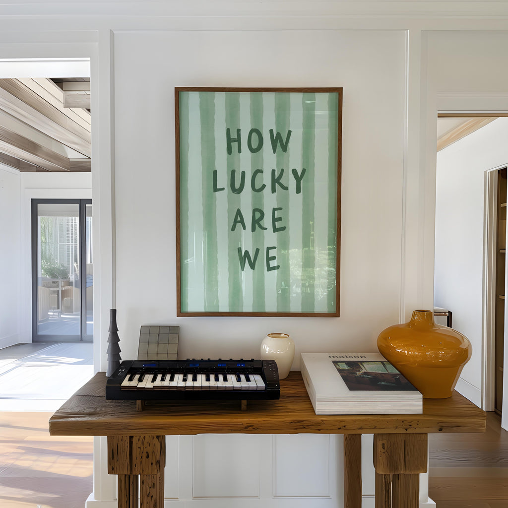 a wooden table with a keyboard, a vase, and a book, along with a framed poster on the wall that reads "HOW LUCKY ARE WE".